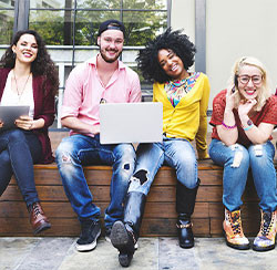 4 young adults sit on ledge with laptop smiling