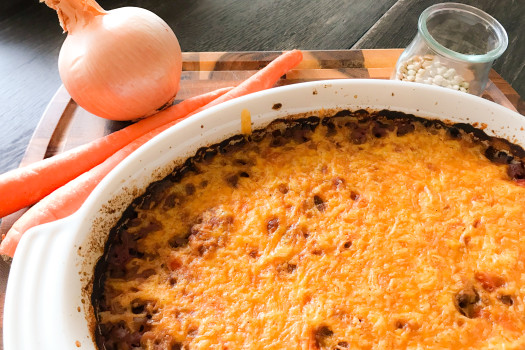 casserole dish of baked lentils with melted cheddar cheese on top with fresh onion and carrots in the background