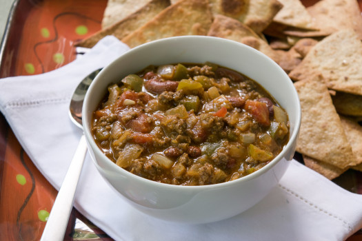 Bowl of beef and bean chili verde with a spoon and pita chips in the background