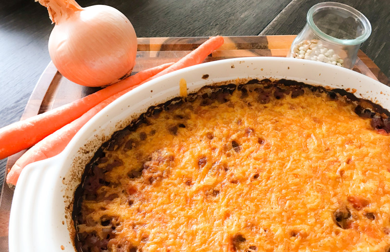 casserole dish of baked lentils with melted cheddar cheese on top with fresh onion and carrots in the background