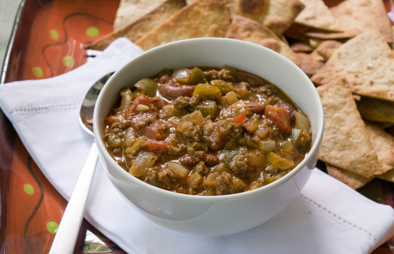 Bowl of beef and bean chili verde with a spoon and pita chips in the background